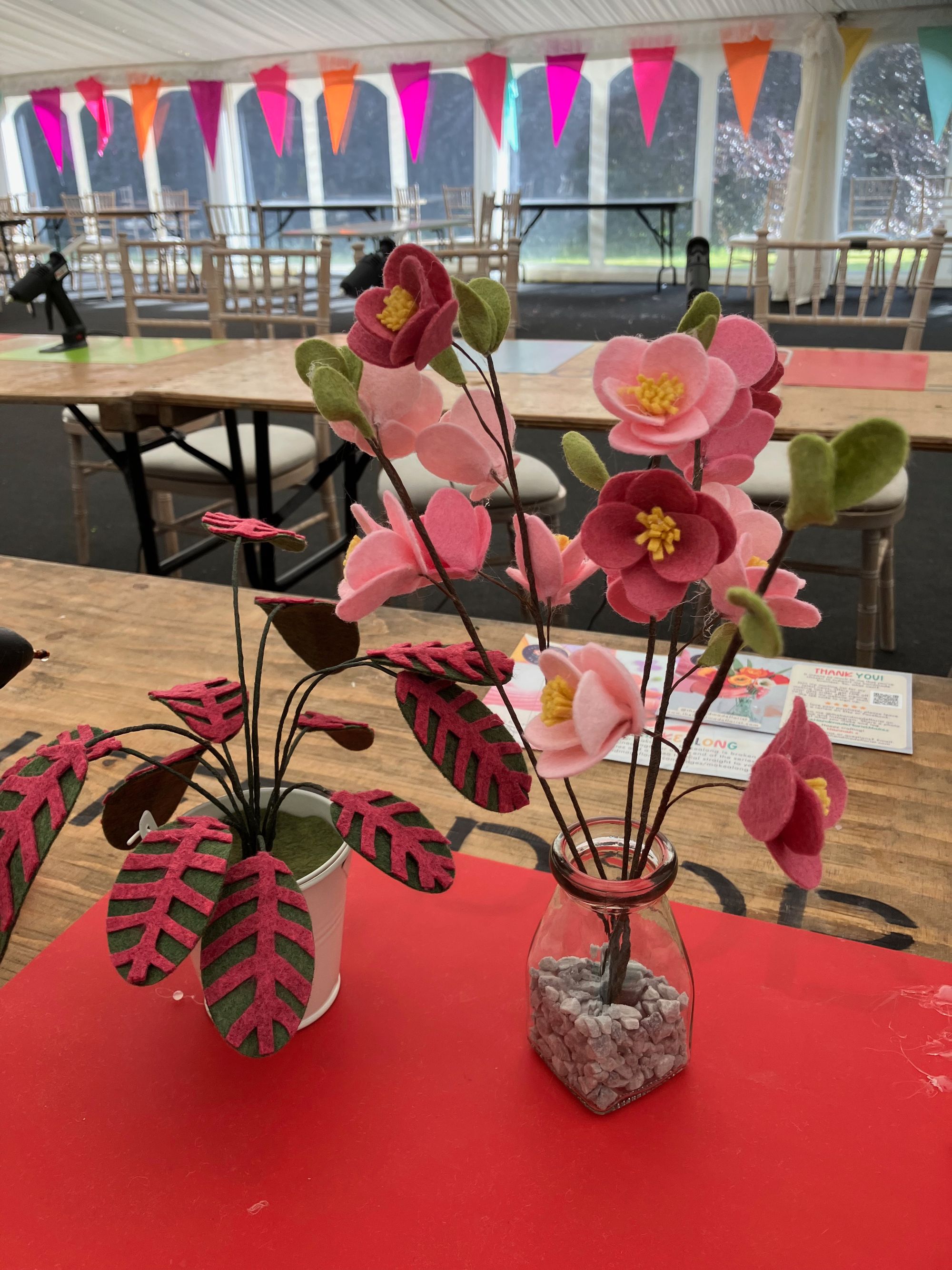 image of felt pink cherry blossoms in a vase and a pink and green felt plant, on a red safety mat on a table in a marquee with pink bunting.